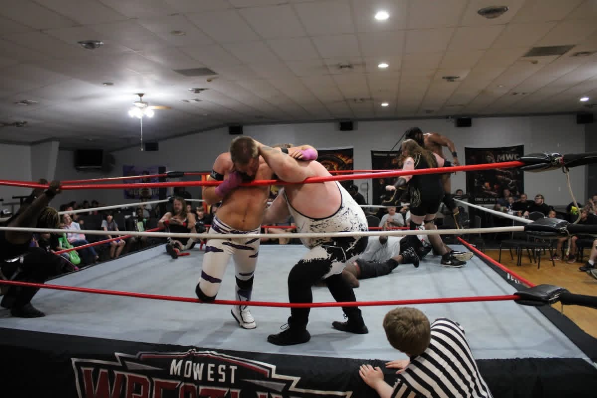 The Apex with purple hand tape grappling with another wrestler near the ropes